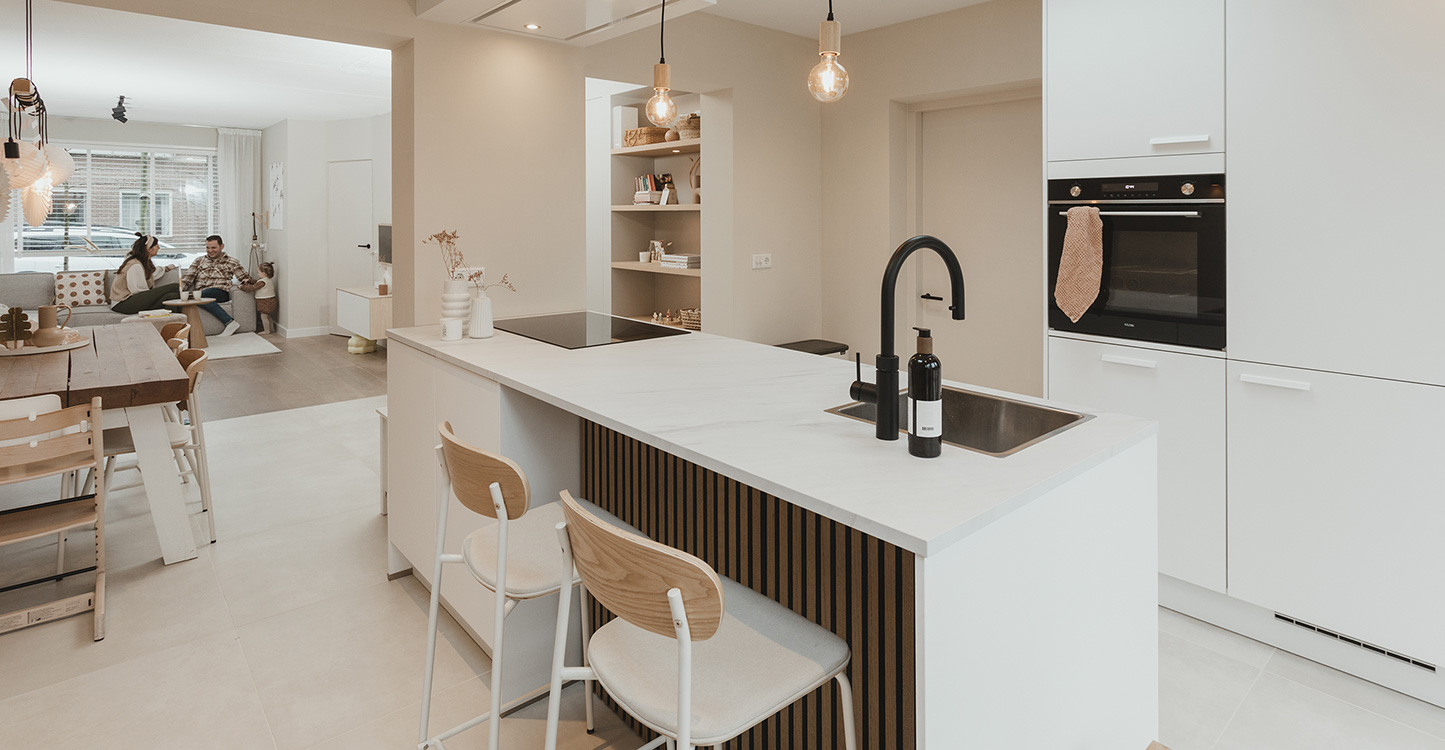 Moderne witte keuken met schiereiland en inductiekookplaat. Oven in de witte hoge kasten. En bar met houtdetail. Achtergrond familie Pieper in de woonkamer